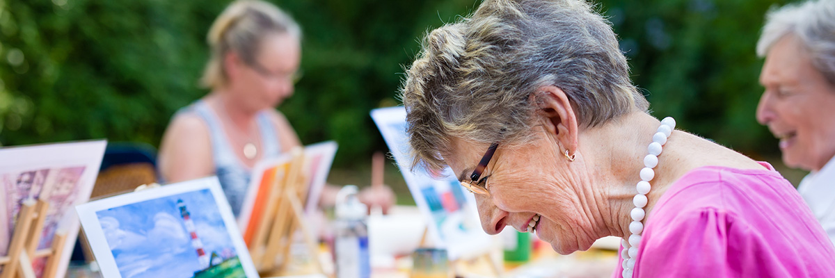 Side view of a happy senior woman smiling while drawing as a recreational activity or therapy outdoors together with the group of retired women